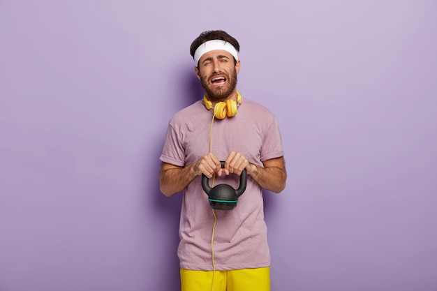 Hard training concept. Sorrowful unhappy young man lifts heavy weight, being demotivated for sport, has no wish to work in gym, wears white headband, casual clothes, listens music during workout