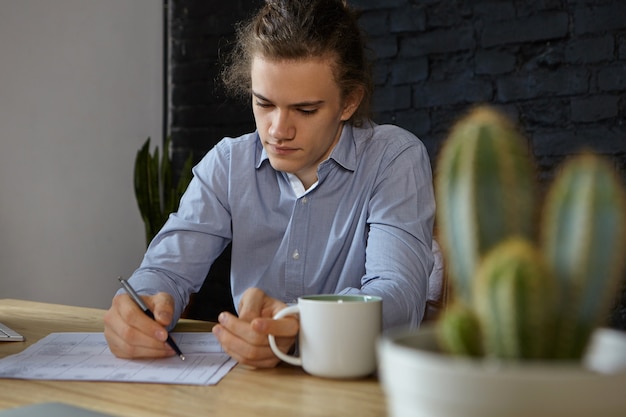 Hardworking concentrated young businessman in blue shirt doing paperwork in his office, having morning coffee. People, job, occupation, profession, business and career concept. Selective focus