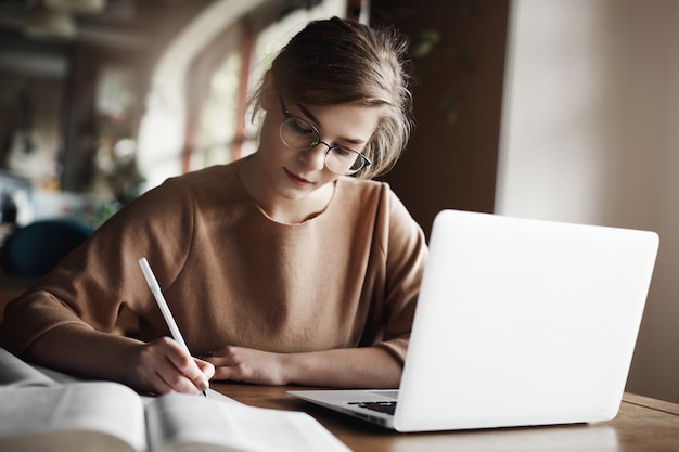 에세이 작성에 집중하고 노트북 근처의 아늑한 카페에 앉아 신중하게 작업하고 메모를 작성하는 데 집중하는 트렌디 한 안경에 열심히 일하는 여성.