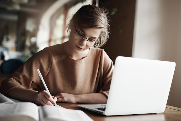 Hardworking focused woman in trendy glasses concentrating on writing essay, sitting in cozy cafe near laptop, working and making notes carefully.