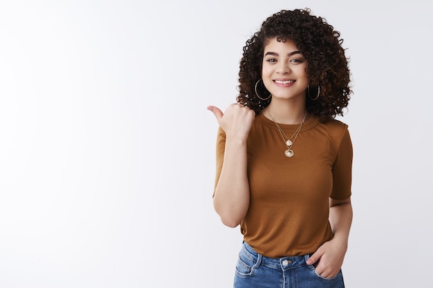 Have you met my friend. Glad good-looking confident stylish female curly dark hair pointing thumb left introduce product smiling happily recommend good promo hold hand jeans pockets assured