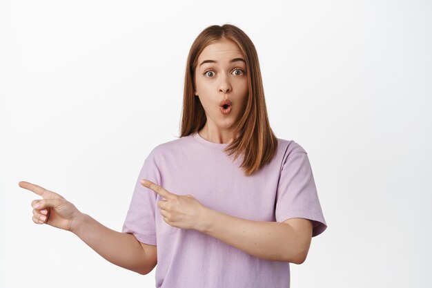 Have you seen it. Image of thrilled blond girl checking out new promo deal, gasp and say wow, stare surprised and excited, pointing finger left at logo, standing against white background.