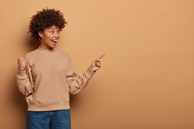 Have you seen it. joyful surprised ethnic girl shows way to awesome new event or promo banner, smiles happily and clenches fist, feels triumph, glad to reach desirable item, isolated on brown wall