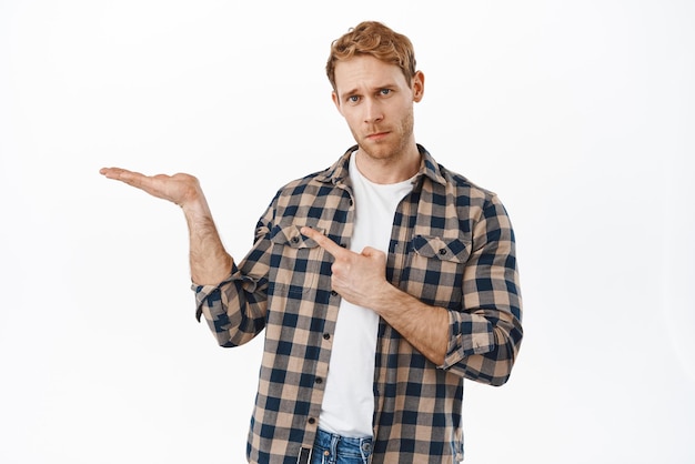 Free photo have you seen this serious redhead man pointing at object on his open hand and looking questioned askign question about an item on palm standing over white background