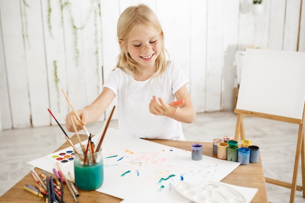 Having fun, joyful, smiling with teeth blonde seven-year-old girl dripping paint over white sheet of paper lying on a table. Creative child having fun, enjoying painting.