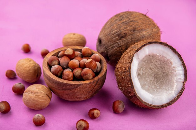 Hazelnuts on a wooden bowl with fresh coconuts and walnuts isolated on pink surface