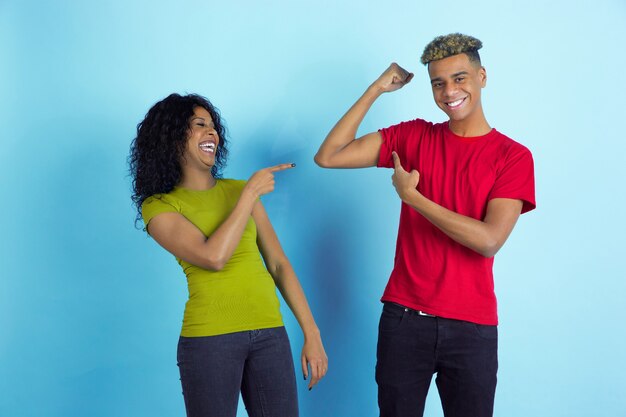 He's strongs, she's laughting. Young emotional african-american beautiful man and woman in colorful clothes on blue background. Concept of human emotions, facial expession, relations, ad, friendship.