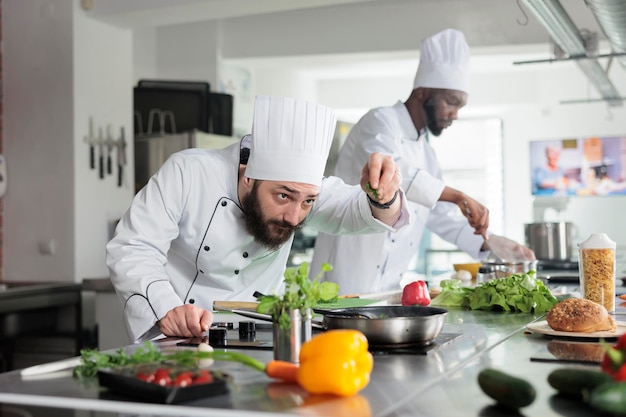 Head chef adding fresh chopped green herbs in pan while cooking gourmet dish in restaurant professional kitchen. Food industry workers preparing delicious meal using organic vegetables.