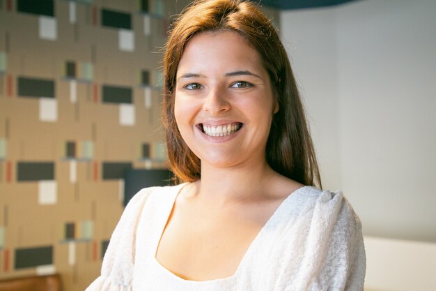 Head shot of happy beautiful young woman posing indoors, looking at camera and smiling