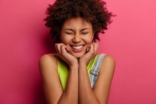 Head shot of positive curly haired woman keeps hands under chin, closes eyes and smiles broadly, enjoys nice time in good company, dressed in fashionable clothes, isolated on pink wall