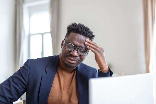 ヘッドショットは、ホームオフィスのコンピューターで作業しているひどい頭痛に苦しんで額に触れている若いアフリカ系アメリカ人の男性を強調しました頭に痛みを伴う感情を持っている欲求不満の混乱した異人種間の男
