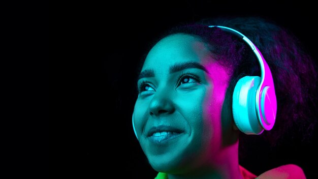 Headphones. African-american woman's portrait isolated on dark studio background in multicolored neon light. Beautiful female model. Concept of human emotions, facial expression, sales, ad, fashion.