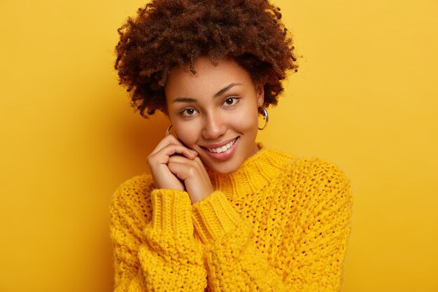 Headshot of adorable romantic woman has pleasant smile, tilts head on hands, has tender look, has dark curly hair, wears cozy winter sweater, isolated over yellow background.