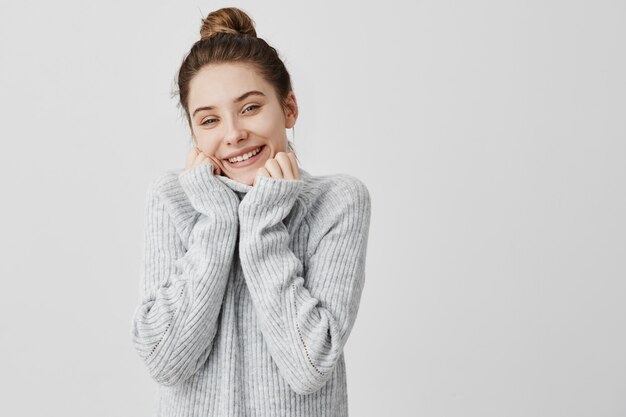 Headshot of adorable smiling woman wrapping up her face in collar of grey sweater. Female artist expressing appeasement and comfort. Feelings concept
