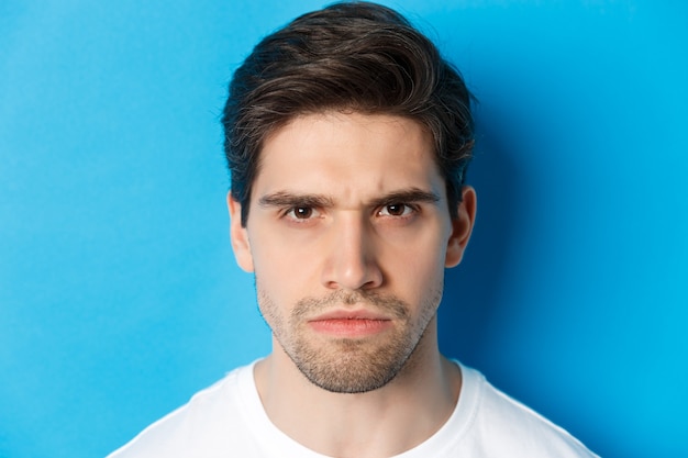 Headshot of angry man frowning, looking disappointed and bothered, standing over blue background