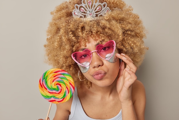 Headshot of attentive curly haired woman applies silver beauty patches under eyes to remove wrinkles wears trendy pink sunglasses holds big multicolored lollipop isolated over grey background