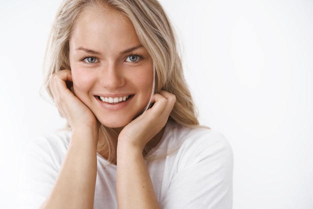 Headshot of attractive adult blond woman with tattoo leaning face on palms and smiling lovely making romantic gazes at camera feeling soft and silk skin applying cosmetics mask skincare products