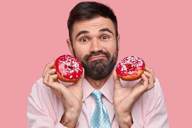 Headshot of attractive bearded man with black hair, has friendly expression, holds red delicious donuts, dressed in formal clothes, models against pink background
