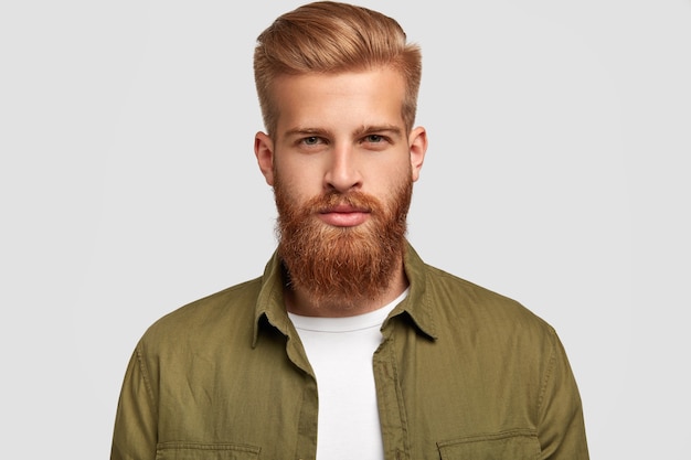 Free photo headshot of attractive bearded man with trendy hairdo, has foxy thick beard and mustache, looks seriously, listens attentively news from interlocutor, isolated over white wall. lifestyle concept