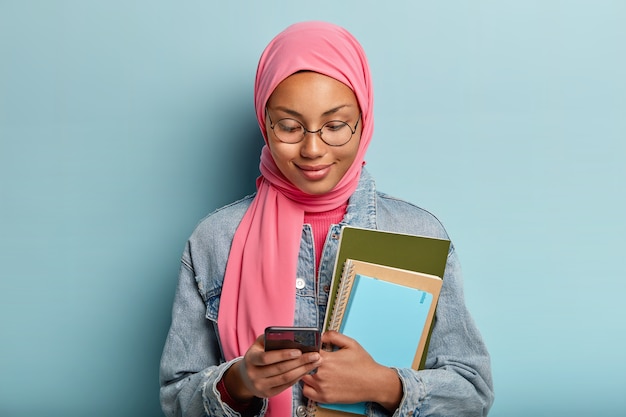 Headshot of attractive mixed race Arabian woman focused in cell phone, works with files on cellular, likes new app, holds spiral notebook, dressed in denim jacket and pink hijab according to religion.