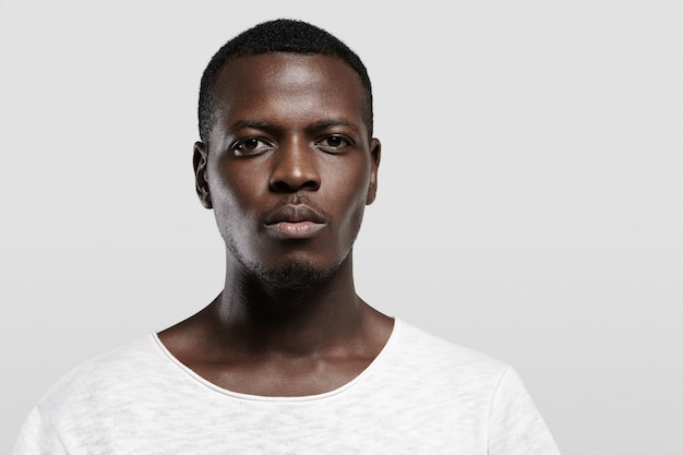 Free photo headshot of attractive serious african student with small beard and moustache, dressed in casual t-shirt  with confident and thougthful expression on his face standing at gray wall