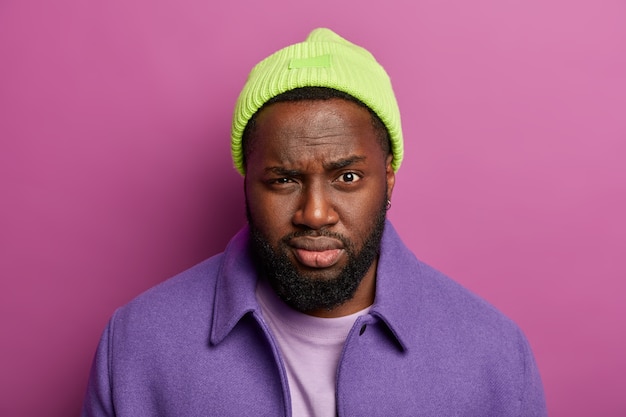 Headshot of attractive serious man with dark skin, full lips, looks mysteriously at camera, wears green hat and purple coat, stands indoor