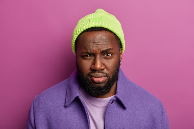 Headshot of attractive serious man with dark skin, full lips, looks mysteriously at camera, wears green hat and purple coat, stands indoor