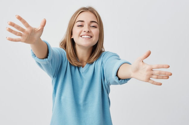 Headshot of attractive smiling young female dressed in blue sweater and outstretching her arms with joy. people, happiness and positive emotions concept.