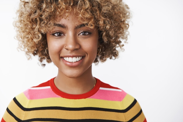 Foto gratuita colpo in testa di giovane donna dalla carnagione scura attraente e dall'aspetto amichevole dall'aspetto gioioso con un taglio di capelli riccio equo in maglione a righe sorridente felice e ottimista alla macchina fotografica sul muro bianco