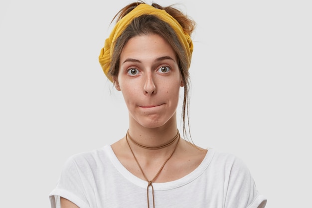 Headshot of attractive woman presses lips together, has confused facial expression, looks , wears yellow headwear on head