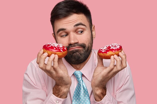 Headshot of attractive young man has puzzled facial expression, holds tasty doughnuts, looks confusingly, has temptation