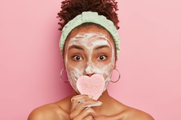 Headshot of attractive young woman with fresh healthy skin, has hygienic procedures during weekend, wears headband, covers mouth by cosmetic sponge, wears round earrings, looks with widely opened eyes