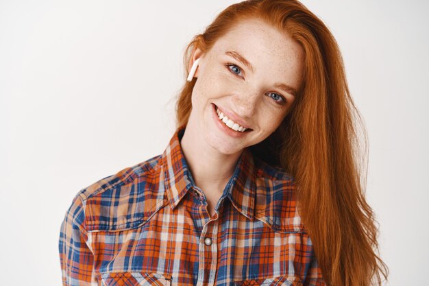 Headshot of attractive young woman with long red hair and blue eyes, listening music in wireless headphones, smiling dreamy at front, white wall