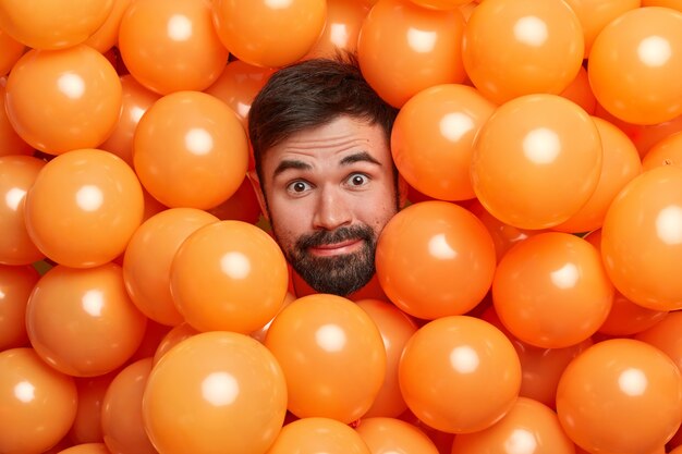 Headshot of bearded adult European man surrounded with inflated orange balloons prepares for party