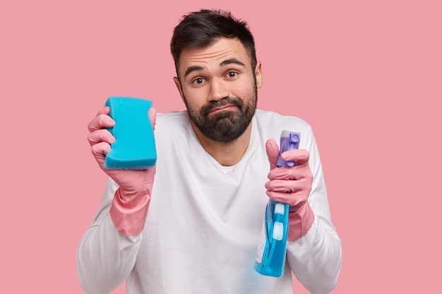 Headshot of bearded young European man has thick beard, wears rubber gloves and white casual jumper, looks with clueless expression