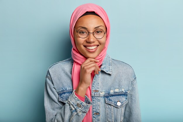 Headshot of beautiful Arabian woman with toothy smile, holds chin, wears round spectacles, wears special traditional clothes