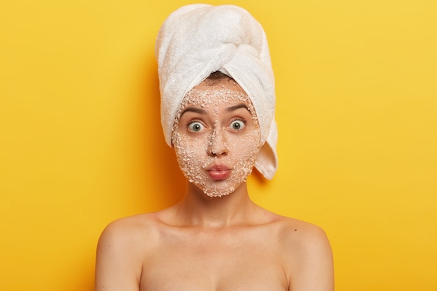 Headshot of beautiful Caucasian woman has widely opened eyes, keeps lips folded, cares about purity and freshness of skin, has surprised face expression, well cared body, takes shower in bathroom