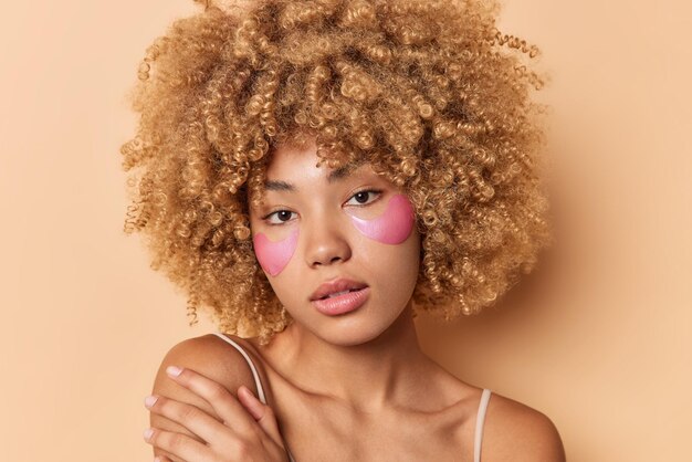 Headshot of beautiful curly haired young woman has soft skin touches shoulder gently looks directly at camera applies pink hydrogel patches under eyes isolated over brown background. Beauty concept