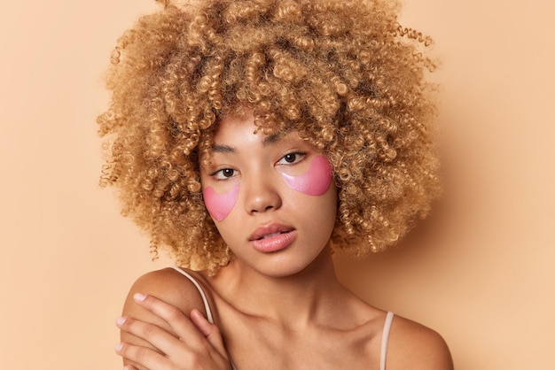 Headshot of beautiful curly haired young woman has soft skin touches shoulder gently looks directly at camera applies pink hydrogel patches under eyes isolated over brown background. Beauty concept