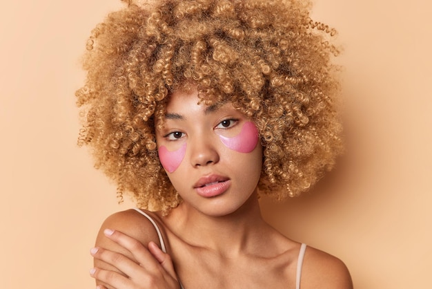 美しい縮れ毛の若い女性のヘッドショットは、柔らかい肌が肩に優しく触れ、カメラを直接見て、茶色の背景の上に分離された目の下にピンクのヒドロゲルパッチを適用します。美容コンセプト