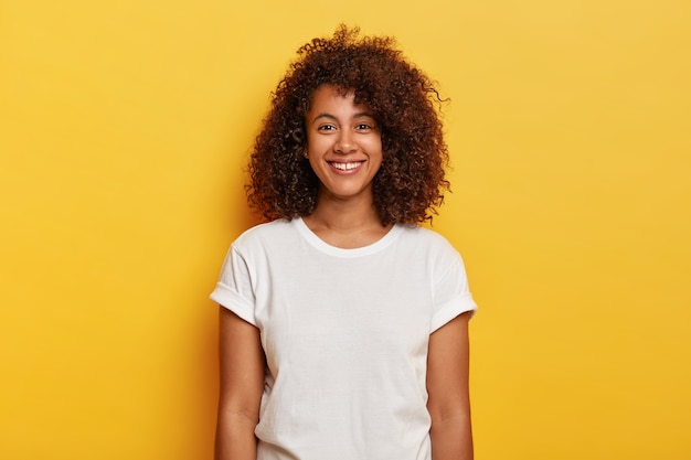 Headshot of beautiful dark skinned curly has pleased expression, rejoices success, enjoys spare time, wears casual t shirt, isolated on yellow wall. People, positive emotions, feelings concept