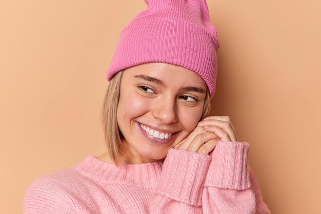 Headshot of beautiful dreamy European woman keeps hands near face smiles happily concentrated away wears pink hat and comfortable jumper has pleasant thoughts isolated over brown background