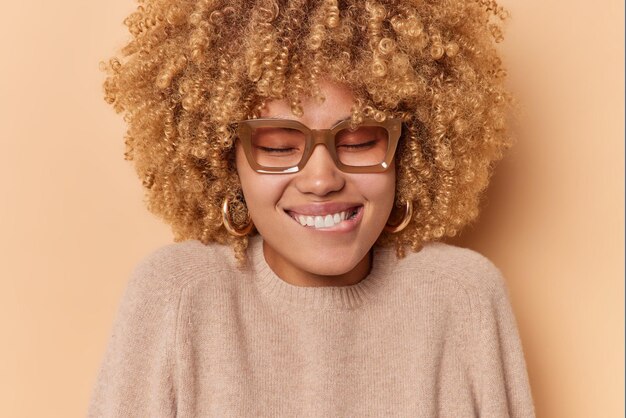 Headshot of beautiful dreamy woman bites lips feels positive stands with closed eyes recalls nice memories wears eyeglasses and sweater isolated over brown studio wall. People emotions feelings