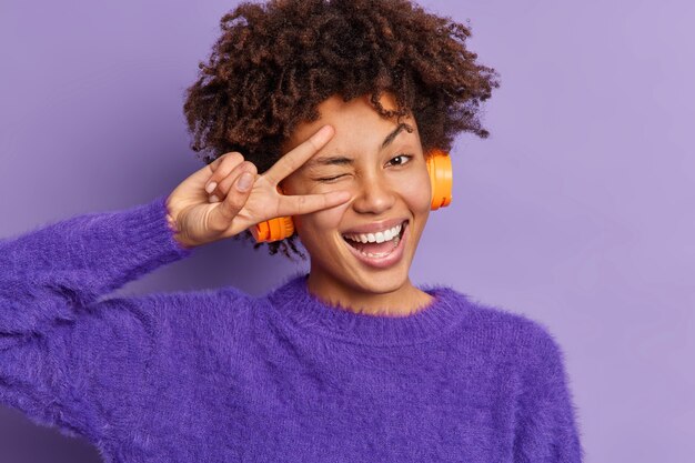 美しい楽しませてくれた暗い肌の女性のヘッドショットは目をまばたきし、平和のジェスチャーを示します勝利のサインを笑顔で広くワイヤレスヘッドフォンで音楽を聴きます暖かい紫色のセーターを着て屋内に立っています