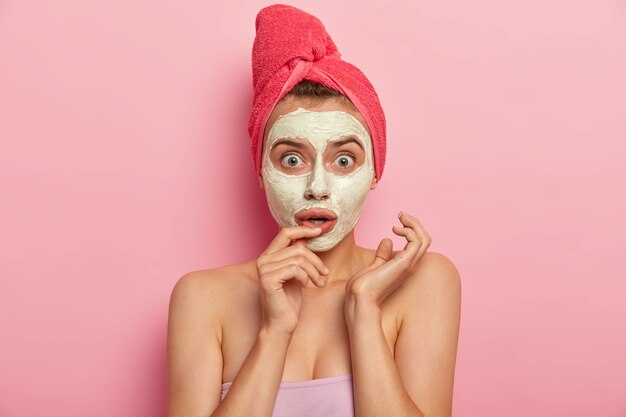 Headshot of beautiful European woman applies natural clay mask, visits spa salon, wears pink soft towel, has stupefied reaction, cares about body, models indoor. Exfoliation, freshness, skin care
