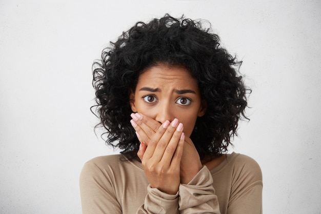 Free photo headshot of beautiful scared young dark-skinned european female with curly hairstyle closing mouth not to scream, feeling frightened and terrified, her eyes and look full of fear and terror