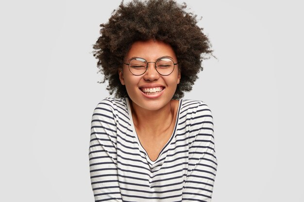 Il primo piano del volto della bella femmina dalla pelle scura divertente sorridente ha un taglio di capelli afro, ride di qualcosa, tiene gli occhi chiusi dal piacere, vestito con un maglione a righe, isolato sopra il muro bianco. felicità