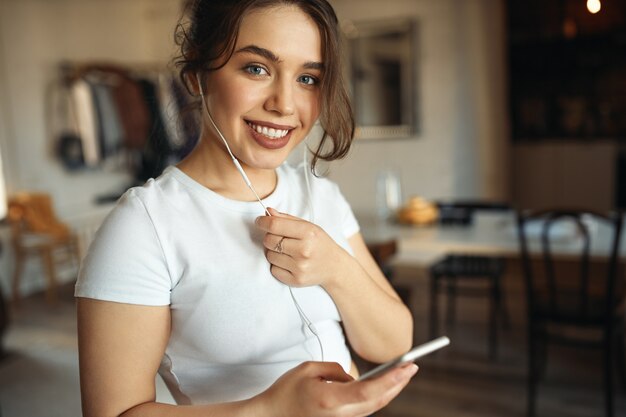 Headshot of beautiful teen girl with chubby cheeks and curvy body posing in living room using wireless internet connection communicating online via audio chat using cell phone and earphones