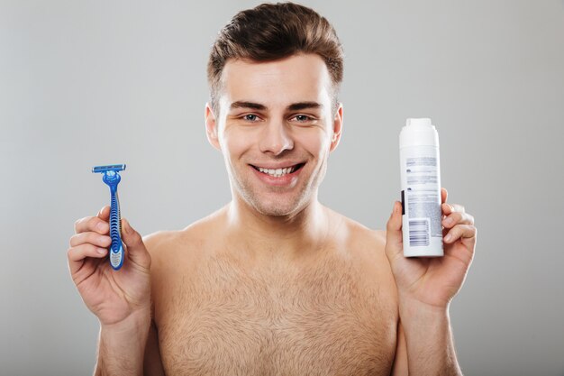 Headshot of brunette happy guy 30s being undressed at bathroom holding razor and shaving cream over grey wall