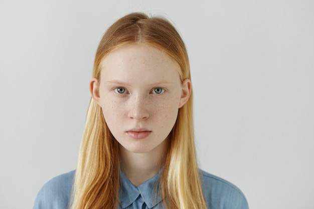 Headshot of Caucasian girl with freckles, blond hair and light eyes dressed in blue school shirt standing against white wall